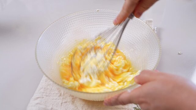 A person whisks beaten egg yolk into soft white curd inside a textured glass bowl, steadying the bowl with one hand on a linen cloth.