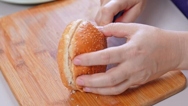 A woman holds a sesame seed bun and cuts it open with a kitchen knife on a wooden board, leaving soft white crumbs. Two halves lie ready for a homemade sandwich in a home kitchen.