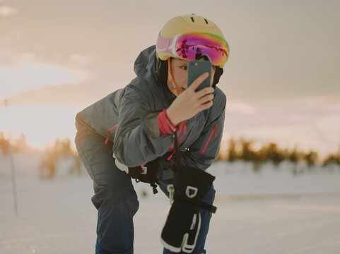 Woman skier in helmet taking a smartphone selfie on a snowy slope at sunset