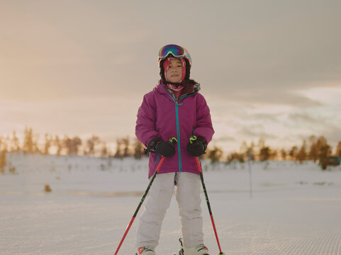 Portrait of a young girl skier with goggles and helmet against a snowy sunset