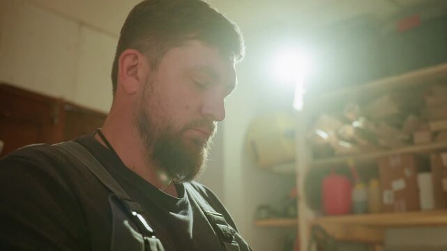 Man in dark shirt wears work apron standing or sitting quietly inside well lit woodworking shop. Workshop shelves hold various wooden planks boxes red container under bright light source