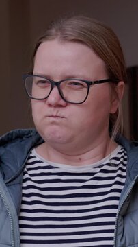 A woman in dark rimmed glasses, a striped top and a blue jacket widens her eyes in surprise, then purses her lips and puffs out her cheeks in a sulking, doubtful reaction indoors at home.