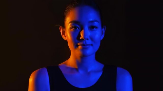 Adult woman raises her gaze from downward to direct eye contact while centered against a black background, with dramatic blue and orange split lighting shaping her face and shoulders