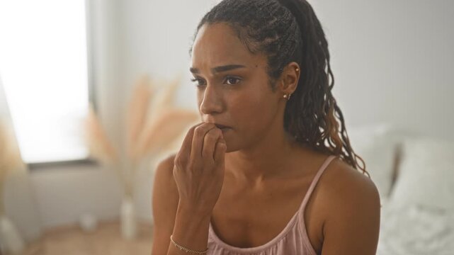 Woman bites fingernails with hand to mouth while seated on a bed in a bright bedroom, wearing tank top and ponytail; anxiety reflection.