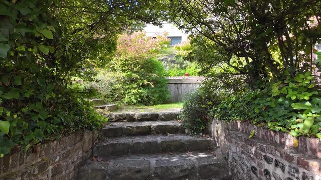 Slow motion pullback reveals a lush garden path, stone steps and wooden gate surrounded by dense vegetation in Veules-les-Roses, Normandy, France