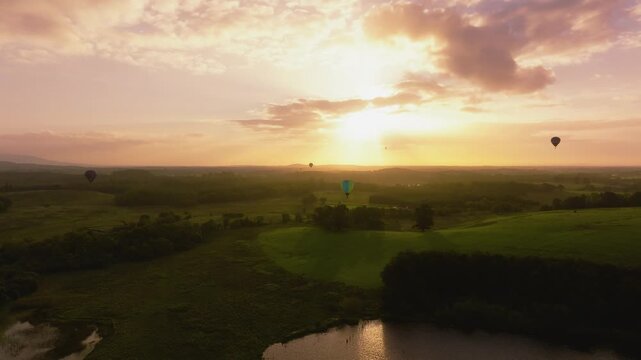 Colorful hot air balloons float above rolling green fields and a small lake at warm sunrise, with soft clouds glowing on the horizon.