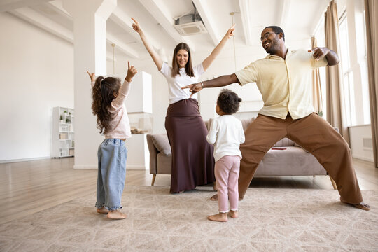 Multiethnic couple with daughters dancing in living room, family having fun at home, spending time together, enjoy active leisure, celebrating, moving to favourite music. Parents and children bonding