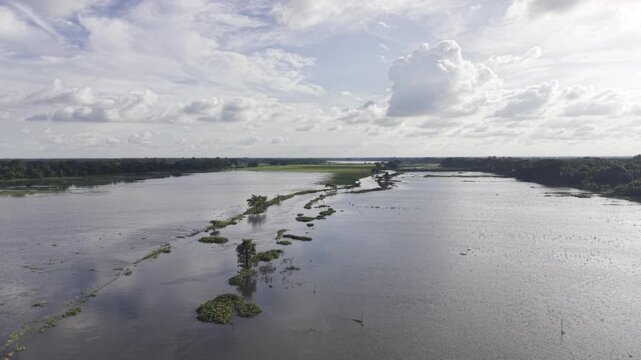 Drone moves forward over monsoon-flooded agricultural land in Kushtia, Bangladesh, revealing a narrow waterway, submerged fields, green grassy village banks, and a blue sky with white clouds.