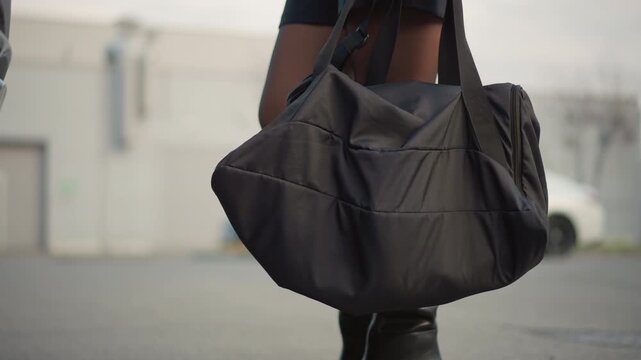 Shows black duffel bag held by person with partial visibility of legs and boots in outdoor parking area. scene is set outdoors on pavement ground with blurred vehicles visible