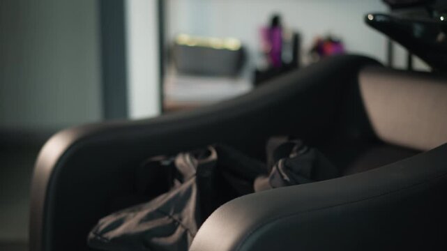 Close up shows dark colored beauty treatment area featuring unoccupied stylist station. padded armrest and part of salon chair are visible with soft lighting creating gentle shadows