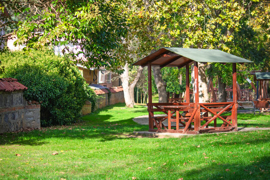 A wooden gazebo on the green grass beneath the trees. Gazebo or pergola in the park or grove for a picnic. Open area, public. Nature. No people, nobody. Horizontal photo.