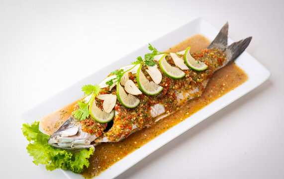 Thai steamed whole sea bass, spicy lime sauce, garlic, chili, and coriander topping on a traditional ceramic plate, authentic exotic seafood dish, steamed seabass with lemon sauce