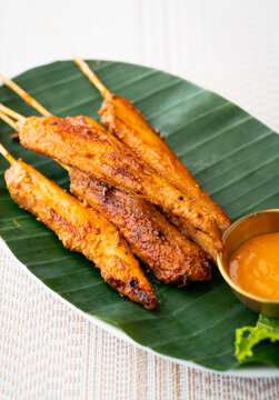 Grilled chicken skewers coated in a golden spicy marinade, served on a green banana leaf with orange dipping sauce on a white plate, photographed close up in bright natural light for restaurant menus.