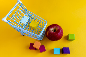 Shopping cart with cubes beside red apple and scattered blocks on yellow background © Irina