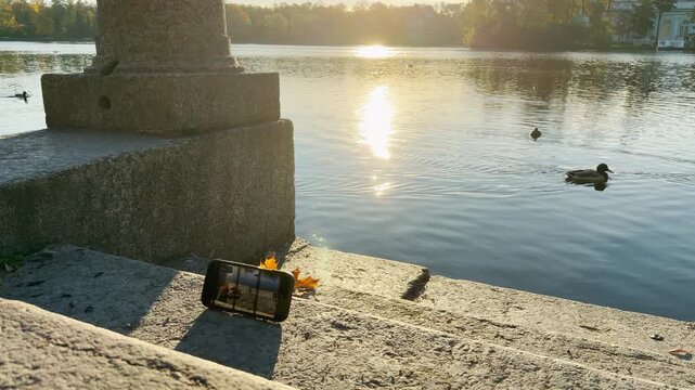 A phone rests on stone steps by a lake. Sunlight glimmers over Catherine Park, Pushkin, Saint Petersburg. Slow motion captures a peaceful and serene atmosphere.