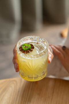 Woman holding a rimmed yellow cocktail glass garnished with a dried lime slice and jalapeno.
