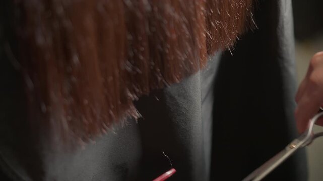Close up shows person long dark brown hair being sectioned and cut by hands holding red comb. Red nail polish is visible on hand with scissors while another pair of hands holds matching red