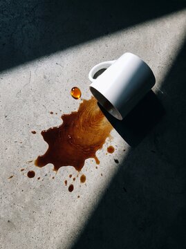 A white ceramic mug lies on its side on a concrete floor with dark coffee spilled next to it, illuminated by a strong diagonal shadow.