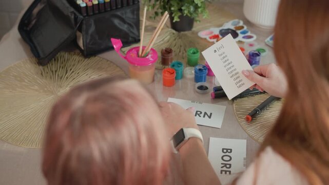Woman holds flashcards with phonetic words like ei face and cake while interacting closely with young girl at table. scene includes colorful paint containers markers brushes in pink cup art