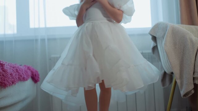 Young girl in white ruffled skirt and shoes moves gracefully indoors her feet visible as girl twirls. Soft natural light fills room with sheer curtains and furniture details like pink fuzzy