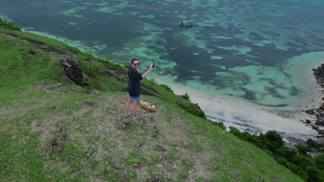 Woman With Dog Taking Selfie On Grassy Cliff Overlooking Turquoise Ocean And Small Boat, Casual Outfit And Sunglasses, Joyful Travel Vibe, Dramatic Shoreline And Panoramic Aerial View,Lombok