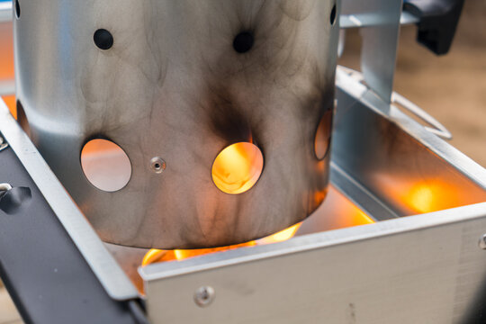 Visible flames rise from a metal charcoal chimney starter placed on a grill, outdoors. The chimney is designed for efficient charcoal lighting, showing fire and smoke around it.