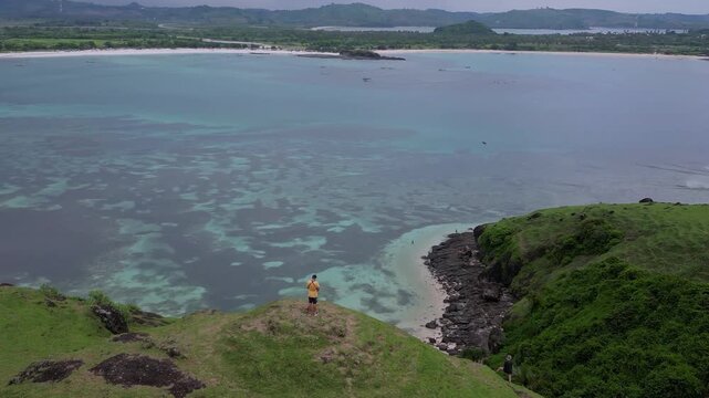 Man standing on cliff overlooking sea,turquoise bay and distant shoreline,lone traveler gazing at coral patterns in shallow water,green grass slope with scattered rocks,Bukit Merese, Lombok