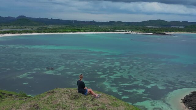 Caucasian woman and dog sitting on cliff overlooking turquoise bay, panoramic aerial view of clear water, sandy cove, green slopes, tranquil mood, gentle breeze, Bukit Merese, Lombok, Tanjung Ann