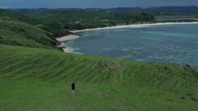 Woman walking hill above turquoise bay, solo hiker pauses to admire coastal vista, green slopes, sandy beach, clear shallow water, distant mountain ridge, Bukit Merese, Lombok, Tanjung Ann