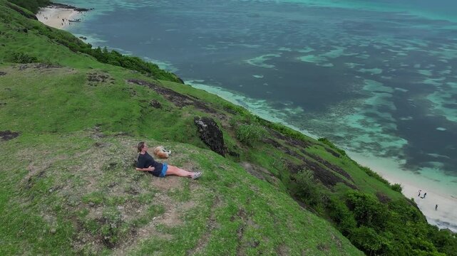 Caucasian woman and dog reclining on grassy ridge above turquoise sea, wide aerial perspective of coral patches and white sand beach, relaxed cuddle, Lombok, Indonesia