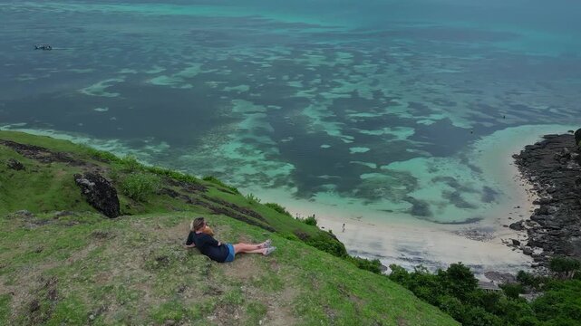Caucasian woman and dog reclining on grassy ridge above turquoise sea, wide aerial perspective of coral patches and white sand beach, relaxed cuddle, Lombok, Indonesia