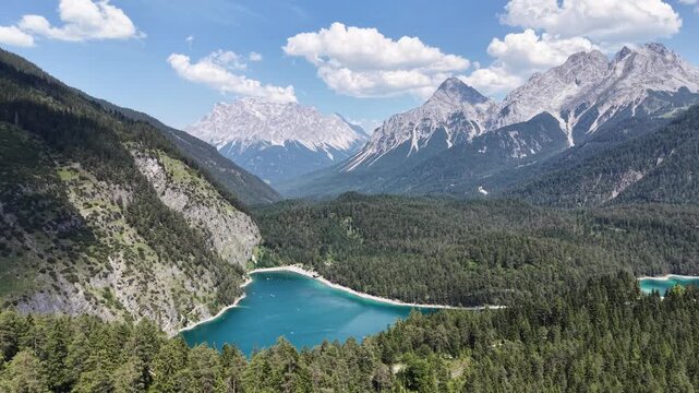 Blindsee turquoise lake nestles in forest below jagged Zugspitze peaks near Biberwier, Austria, aerial drone flies toward clear alpine water.