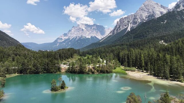 Weissensee emerald lake with tiny island sits below Zugspitze peak near Biberwier, Austria, aerial drone pulls back over alps and forest.