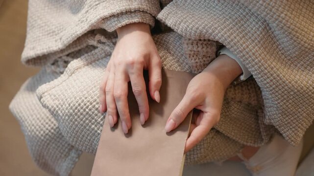 Close up shows two female adult sized hand with long nails gently placed over unopened sealed paper envelope. scene is softly lit emphasizing texture of textured beige fabric and smooth skin