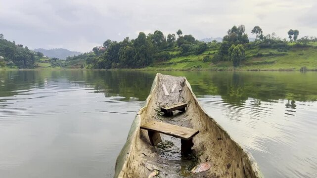 Traditional wooden dugout canoe viewed from the bow on Lake Mutanda, Uganda, with calm water, green cultivated hills and shoreline vegetation surrounding the volcanic lake, first person POV.