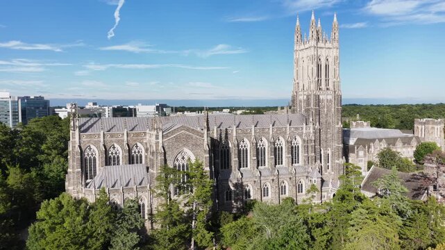 A close orbit of Duke Chapel shows its buttressed nave and arched windows in Durham, North Carolina, campus paths and lawns spreading beneath the tower. Aerial view