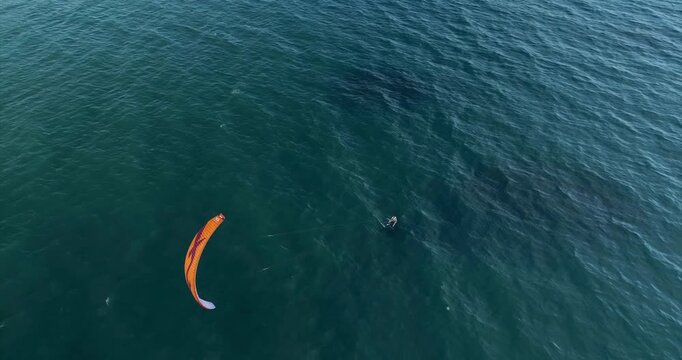 An orange kite sweeps over Dark Teal Cabarete Water from Above as its kitesurfer rides below and leaves a short wake while the drone looks steeply down at the open sea and shifting kite position