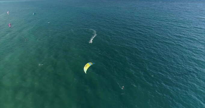 A kitesurfer under a yellow kite cuts a broad curved wake across green blue water while other small riders remain farther out and the drone keeps the moving kite and ocean pattern in view