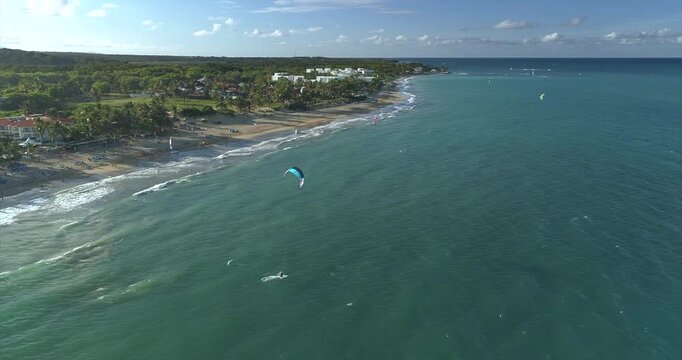A kitesurfer with a blue kite crosses shallow green water beside the Cabarete shoreline while small breakers trace the beach and other distant kites appear along the tropical coast