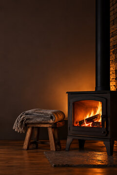 Black wood burning stove with glowing flames and a folded wool blanket on a rustic stool, dark warm evening, vertical with copy space