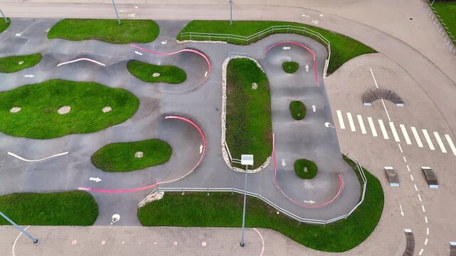 Oblique drone view of a pump track junction with tight bermed turns, grassy islands, safety rails and an adjacent zebra crossing.