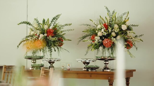 Two tall arrangements of orange and white flowers with eucalyptus stand in glass vases atop silver pedestals, beside small cake stands on a wooden wedding table.