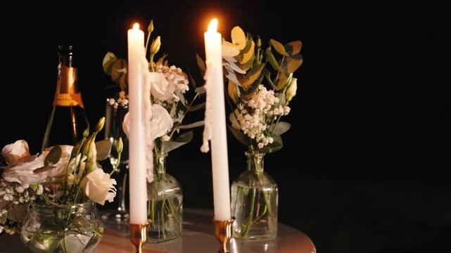 Two tall white candles burn in copper holders among small glass vases of lisianthus and gypsophila, beside a champagne bottle on the table at night.