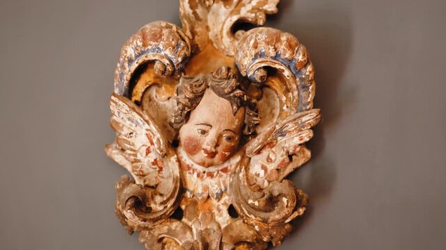 Antique carved wooden cherub head with painted face, gilded scrolls and wings, hanging as decoration on a grey wall.