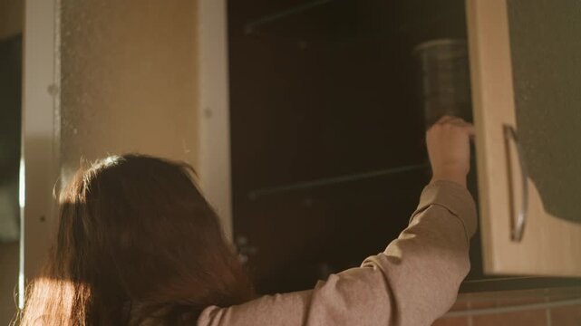 Woman stores green seed filled jars on high kitchen shelves. Woman reaches up to place containers inside cabinets organizing her pantry with methodical movements. Warm sunlight streams