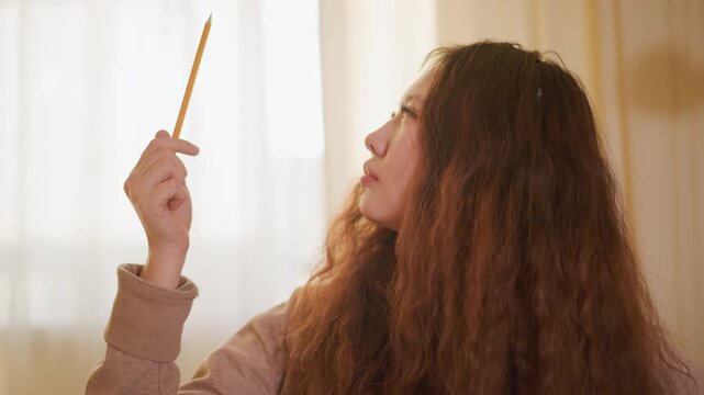 Woman with long hair examines yellow pencil. Soft natural daylight fills room through sheer curtains. scene is calm and focused on her hand holding object