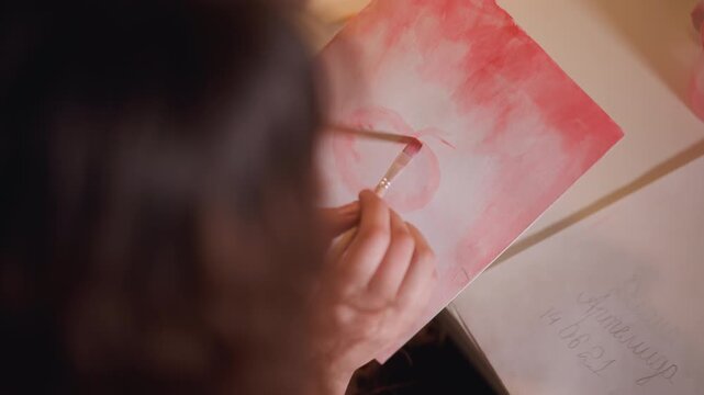 Woman hand holding paintbrush applies soft pink and white wash to blank sheet. Red hue spreads across page as woman works with another sketch partially visible nearby. Warm lighting