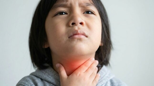 Young Asian child frowning with a sore throat, hand pressed to neck with red inflamed glow, gray hoodie against a plain white wall