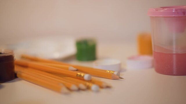 Yellow pencils and paint containers on table. Soft lighting creates gentle shadows from objects. Background remains out of focus emphasizing art supplies in foreground