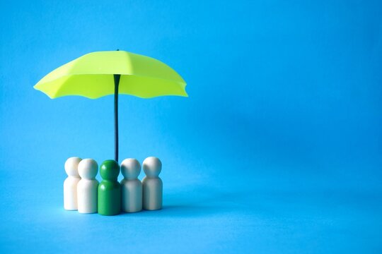 Wooden peg figures with a green leader figure standing under a small yellow umbrella against a vibrant blue background, representing leadership, team protection, and group insurance concepts.